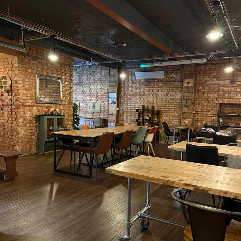 Several long wood-topped tables surrounded by chairs, on a brown floor and with a brick wall, shelves and a mirror in the background, in the first floor at Understated coffee shop. 